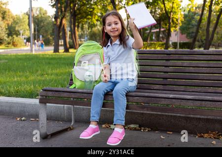 Ein kleines verspieltes Mädchen mit Rucksack, zeigt ein offenes Notizbuch in den Händen, sitzt auf einer Parkbank. Stockfoto