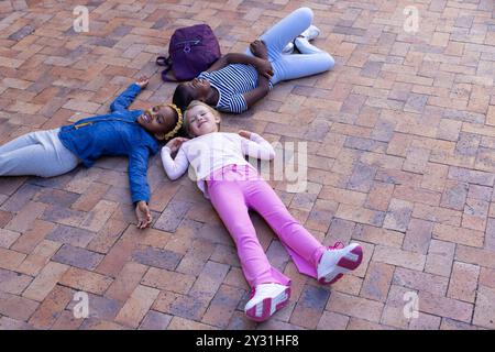 Auf Backsteinpflaster liegend, drei multirassische Mädchen, die sich mit dem Rucksack in der Schule in der Nähe entspannen Stockfoto