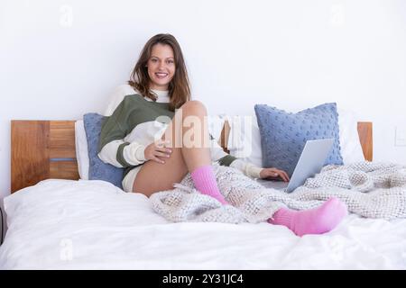 Zu Hause, entspannend im Bett, Frau mit Laptop und lächelnd, mit gemütlichen Socken Stockfoto