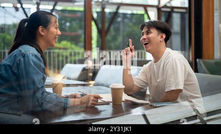 Zwei fröhliche junge asiatische Studenten, ein Mann und eine Frau, unterhalten sich in einem Café. Mit Begeisterung heben sie die Finger, Stockfoto