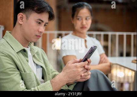 Ein ernsthafter junger Asiate scrollt auf seinem Handy und ignoriert seine launische Freundin, während sie zusammen in einem Café sitzen und eine Beziehung haben Stockfoto