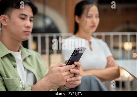 Ein ernsthafter junger Asiate scrollt auf seinem Handy und ignoriert seine launische Freundin, während sie zusammen in einem Café sitzen und eine Beziehung haben Stockfoto