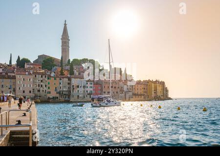 Die Sonne strahlt ein warmes Leuchten über Rovinj Kroatien und beleuchtet farbenfrohe Gebäude entlang des Ufers. Ein Segelboot schwimmt sanft auf dem klaren Wasser, Inv Stockfoto