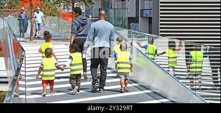 Rückansicht zwei Erwachsene Betreuer gehen auf erhöhtem öffentlichen Gehweg fünf kleine Kinder halten Hände gut sichtbare Westen spiegeln sich in der Glasbalustrade London UK wider Stockfoto