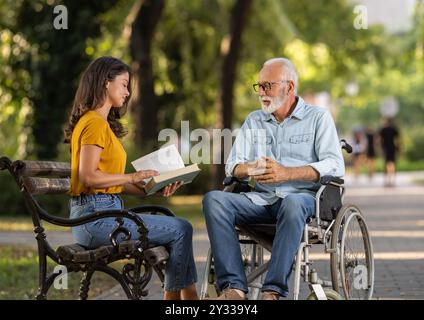 Junge Pflegerin liest einem Seniorenmann im Rollstuhl im Park ein Buch Stockfoto