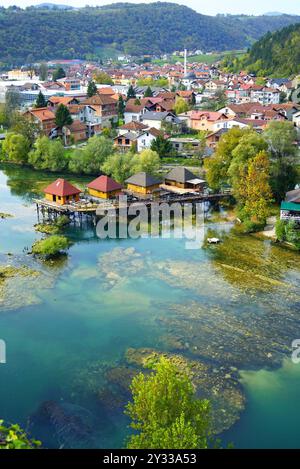 Blick aus der Vogelperspektive auf das Stadtzentrum von Bosanska Krupa, Bosnien und Herzegowina: Der farbenfrohe Fluss Una, traditionelle Pfahlhäuser und flache Gebäude Stockfoto