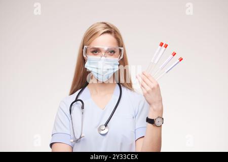 Eine medizinische Fachkraft, die eine Maske und eine Schutzbrille trägt und Reagenzgläser mit roten Kappen hält. Sie hat lange Haare und ein Stethoskop um h Stockfoto