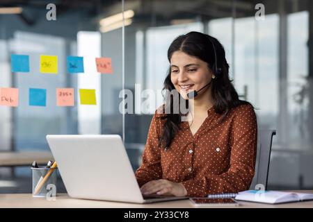 Junge schöne Online-Support-Mitarbeiterin am Arbeitsplatz, hispanische Frau, die Headset-Telefon für Videoanrufe verwendet, Geschäftsfrau, die auf Computertastatur smil schreibt Stockfoto