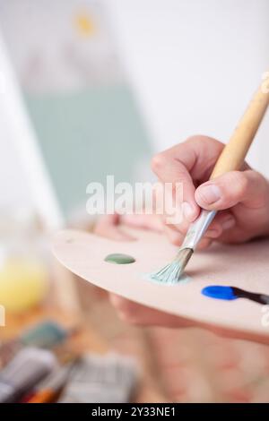 Nahaufnahme einer Person, die mit einem Pinsel Farbe aus der Palette eines Malers nimmt. Im Hintergrund die Leinwand mit dem Bildmaterial. Stockfoto