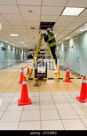 Ein Mann in einer gelben Weste steht auf einer Leiter im Flur. Im Hintergrund befinden sich orangefarbene Kegel. Jorge Chavez International Airport Stockfoto