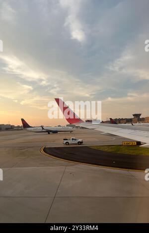 Ein rot-weißes Flugzeug parkt auf dem Asphalt neben einem weißen Auto. Der Himmel ist bewölkt und die Sonne untergeht. Hartsfield Jackson Atlanta International Stockfoto