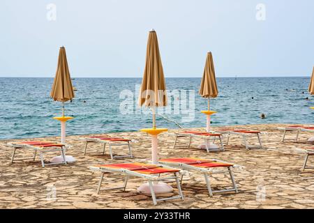 Porec, Istrien, Kroatien - 26. August 2024: Zusammenklappbare Sonnenschirme und leere Liegestühle am Strand von Porec, Kroatien, mit Blick auf das ruhige blaue Meer *** Zugeklappte Sonnenschirme und leere Liegestühle am Strand von Porec, Kroatien, mit Blick auf das ruhige, blaue Meer Stockfoto