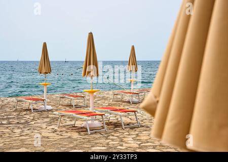 Porec, Istrien, Kroatien - 26. August 2024: Zusammenklappbare Sonnenschirme und leere Liegestühle am Strand von Porec, Kroatien, mit Blick auf das ruhige blaue Meer *** Zugeklappte Sonnenschirme und leere Liegestühle am Strand von Porec, Kroatien, mit Blick auf das ruhige, blaue Meer Stockfoto