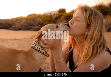 Junge Erwachsene kaukasische Frau küsst ihren Whippet-Hund am Strand bei Sonnenuntergang, liebevoller Moment im goldenen Licht eines Sommernachmittags, Closeup Stockfoto