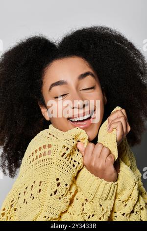 Eine strahlende junge Frau lächelt mühelos, während sie ihren gemütlichen gelben Pullover in einem ruhigen Studio umarmt. Stockfoto