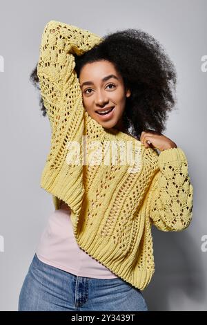 Eine junge afroamerikanische Frau mit lockigen Haaren spielt mit ihrem Pullover und lächelt strahlend in einem Studio. Stockfoto