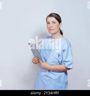 Porträt einer sternförmig aussehenden Schwester mittleren Alters in einem blauen Gestrüpp mit einem Klemmbrett auf neutralem grauem oder grauem Hintergrund. Stockfoto