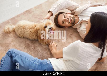 Fröhliches Paar, das auf dem Boden liegt, mit Maltipoo Hund zu Hause spielt und eine fröhliche gemeinsame Zeit genießt. Stockfoto
