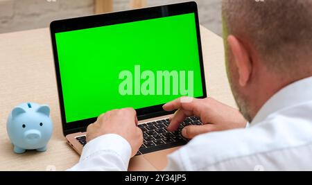 Ein Mann, der einen leeren Laptop mit grünem Bildschirm neben der Schweinebank benutzt. Online-Banking und -Buchhaltung Stockfoto