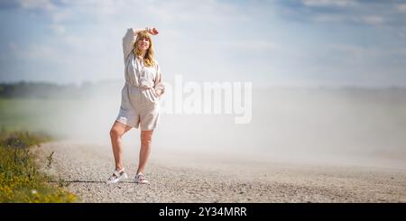 Frau steht auf staubiger Landstraße unter hellem Sommerhimmel. Sie trägt einen leichten Leinen-Strampelanzug und Sandalen und lächelt, während sie ihre Hand auf dem Kopf hält. Staub ri Stockfoto