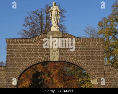 Backsteinbogen mit Statue und herbstlichen Bäumen unter blauem Himmel, Burlo, münsterland, deutschland Stockfoto