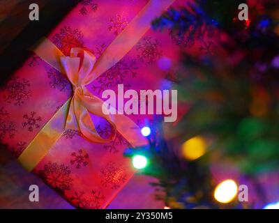 Ein Weihnachtsgeschenk in roter Verpackung liegt unter einem geschmückten Weihnachtsbaum mit bunten Lichtern. Konzept für Weihnachten und Neujahrsfeier. Hintergrund W Stockfoto