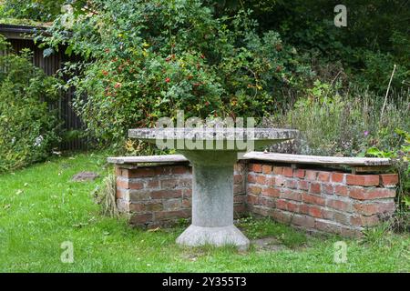 Steintisch und Backsteinbank zwischen Kräutern, einheimischen Pflanzen und wilden Rosenbüschen in einem umweltfreundlichen natürlichen Landgarten, idyllischer Sitzplatz, Kopie Stockfoto