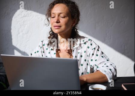 Eine Frau sitzt an einem Tisch und arbeitet an einem Laptop, in natürlichem Licht. Sie genießt eine Tasse Kaffee und vermittelt ein Gefühl von Produktivität und Entspannung. Stockfoto