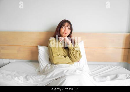 Eine nachdenkliche junge asiatische Frau schaut zur Seite und hält ihr Kinn, während sie auf dem Bett sitzt. Stockfoto