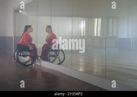 Schöne langhaarige Frau mit Behinderung in einem roten Ballsaal-Kleid, reflektiert im Spiegel im leeren Tanzraum, Seitenansicht. Para tanzen Stockfoto