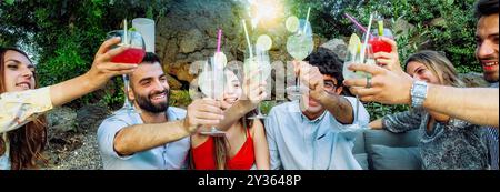 Fröhliche, vielfältige Gruppe junger Freunde, die bei einer Sommerparty im Freien mit bunten Cocktails toasten. Fröhliche Feier in natürlicher Umgebung mit Getränken und Stockfoto