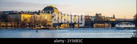 An einem Herbstmorgen zeigt der Moldau-Damm in Prag das in warmes Licht getauchte Tschechische Nationaltheater. Der ruhige Fluss spiegelt die umliegende historische Architektur wider. Stockfoto