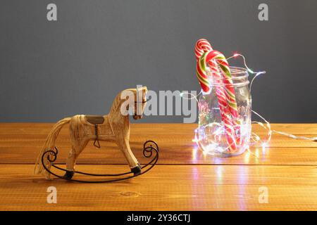 Weihnachtsdekoration - kleines Holzspielzeug Schaukelpferd und Karamell Neujahrsbonbons in einem Glas umschlungsvoll mit einer leuchtenden festlichen Girlande auf einem Tisch Stockfoto