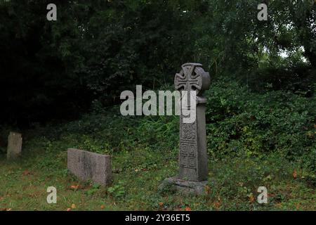 Rowner Lane, Gosport, Hampshire, England. 6. September 2024. Ein keltisches Kreuz und andere Grabsteine in einer dunklen Gegend auf dem Friedhof. Im Domesday Book wird erwähnt, dass Teile der St. Mary's Church in Rowner Lane, Gosport, auf 1.000 Jahre alt sind. Dieses Foto ist eines einer Serie, die ich kürzlich bei einer selbstgeführten Tour durch die Kirche während der Gosport Heritage Open Days gemacht habe. Stockfoto