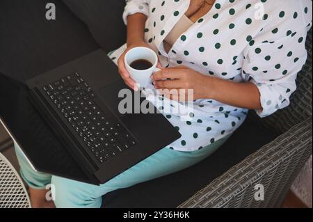Eine Frau genießt eine Kaffeepause, während sie an ihrem Laptop arbeitet, sitzt bequem in einem Korbstuhl und trägt ein stylisches Polka Dot-Shirt. Stockfoto