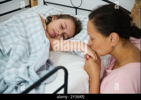 Die Mutter küsst liebevoll die Hand ihrer Tochter, während sie im Bett liegt, was für Fürsorge und Zuneigung steht. Fängt familiäre Wärme und pflegenden Tragekomfort ein. Stockfoto