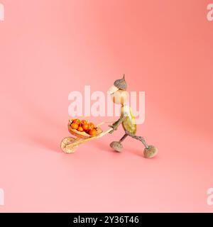 Herbstfigur aus Eicheln, die eine Schubkarre aus Beeren drücken, handgefertigte Konzeptkunst, DIY-Projekt, saisonale Dekoration, kreative Kinderaktivitäten Stockfoto