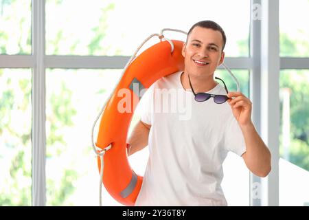 Glücklicher männlicher Rettungsschwimmer mit Ringboje und Sonnenbrille in der Nähe des Fensters im Pool Stockfoto