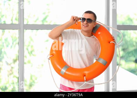 Glücklicher männlicher Rettungsschwimmer mit Ringboje und Sonnenbrille in der Nähe des Fensters im Pool Stockfoto