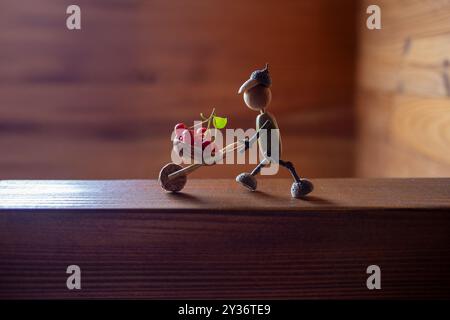Handgefertigte Herbstfigur aus Eicheln mit Zweigbeinen, Eichelkappenkopf, Schubkarre mit Beeren gefüllt, blauer Himmel Hintergrund, ideal für DIY-Handwerk, Stockfoto