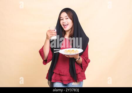 Schöne elegante asiatische Frau in rotem Hemd, die einen Hijab trägt und glücklich lächelt, ein Glas Milch hält und einen Teller mit Dimsum (chinesisches Essen) trägt Stockfoto