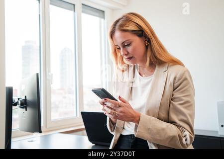 Geschäftsfrau schreibt und sendet E-Mails mit einem Mobiltelefon in ihrem Büro. Bürofrau, die ein Mobiltelefon bei der Arbeit benutzt weibliche Mitarbeiterin oder Sekretärin, die am Arbeitsplatz steht und ein Smartphone ansieht. Hochwertige Fotos Stockfoto