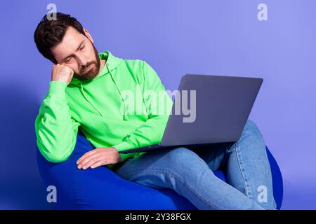 Foto des attraktiven jungen Mannes sitzen Sitzsack halten Netbook müde schlafend gekleidet stilvolle grüne Kleidung isoliert auf violettem Hintergrund Stockfoto
