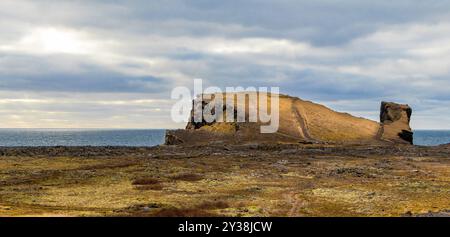 Ein felsiger Hügel mit Blick auf ein Gewässer. Halbinsel Reykjanes. Der Himmel ist bewölkt und die Sonne scheint durch die Wolken Stockfoto