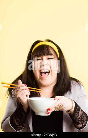 Happy, Frau und Essstäbchen im Studio für Essen, Ernährung und Lachen auf gelbem Hintergrund. Weibliche Person, plus Größe und Schüssel für Ballaststoffe oder gesund Stockfoto