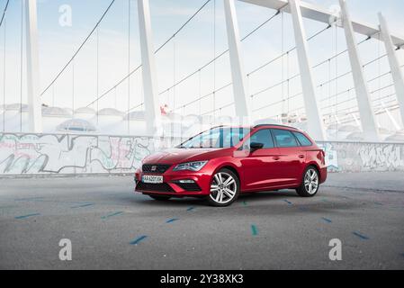 Red Leon Cupra parkte vor dem modernen Stadion. Vorderansicht des sportlichen SITZWAGENS bei Sonnenaufgang, der auf dem großen Asphaltparkplatz geparkt ist. Stockfoto