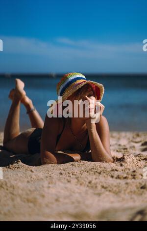 Eine Frau in Badeanzug und mehrfarbigem Hut liegt am Strand. Stockfoto