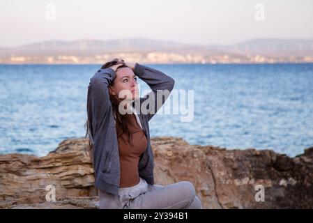Junge, traurige, schöne Frau, die an einem felsigen Strand sitzt und aufblickt Stockfoto