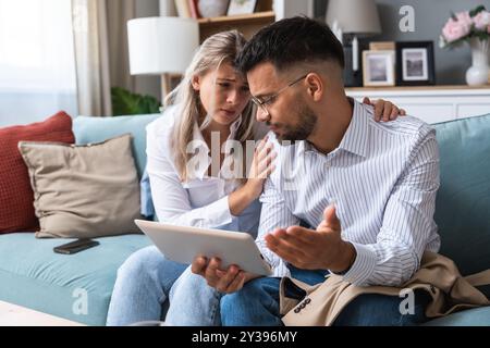 Eine junge Geschäftsfrau tröstet ihren Mann zu Hause, während er negative Statistiken über den Handel seines Unternehmens auf einem digitalen Tablet liest. Zusammengehörigkeit Stockfoto