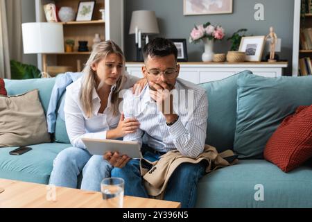 Eine junge Geschäftsfrau tröstet ihren Mann zu Hause, während er negative Statistiken über den Handel seines Unternehmens auf einem digitalen Tablet liest. Zusammengehörigkeit Stockfoto